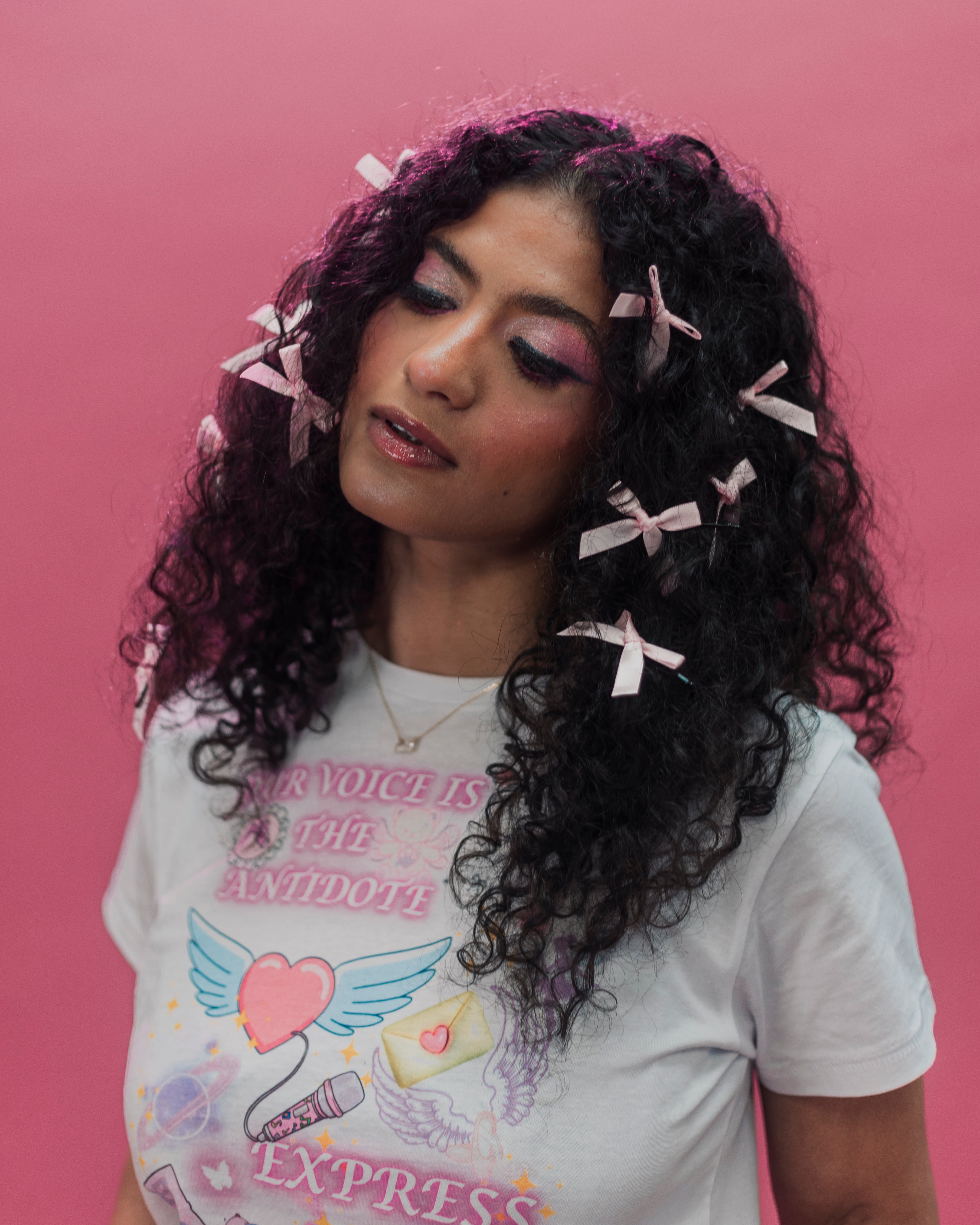 A woman with long, curly dark hair adorned with small pink bows, wearing a white graphic t-shirt with pink text and illustrations, set against a solid pink background.