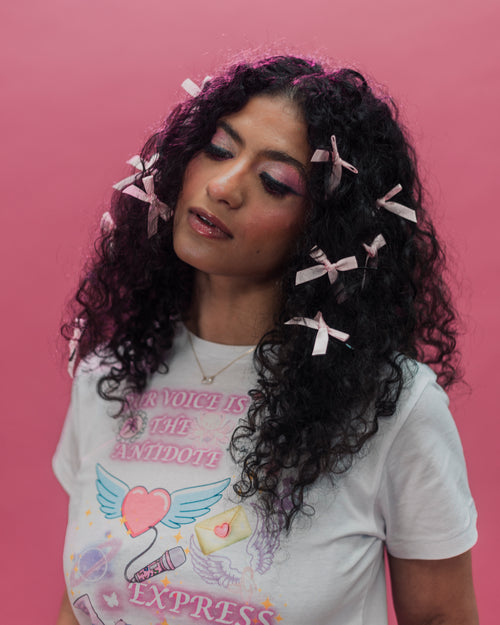 A woman with long, curly dark hair adorned with small pink bows, wearing a white graphic t-shirt with pink text and illustrations, set against a solid pink background.