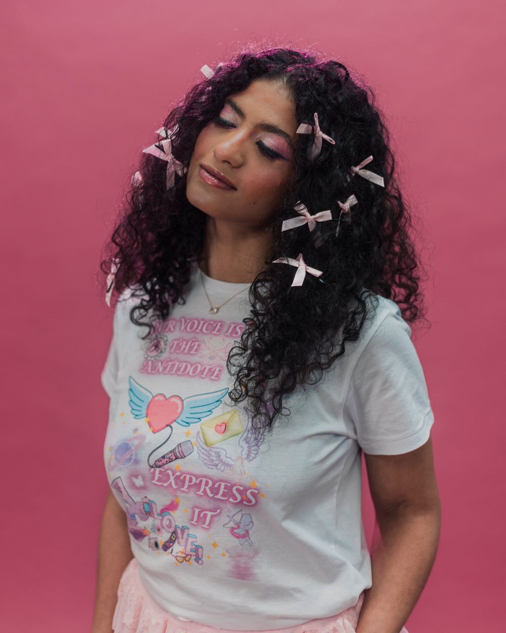A woman with long, curly black hair adorned with pink ribbon clips, wearing a white graphic t-shirt with a colorful angel design and the text 'OUR VOICE IS THE AN ANTIDOTE' and 'EXPRESS IT'. She is posed against a solid pink background.