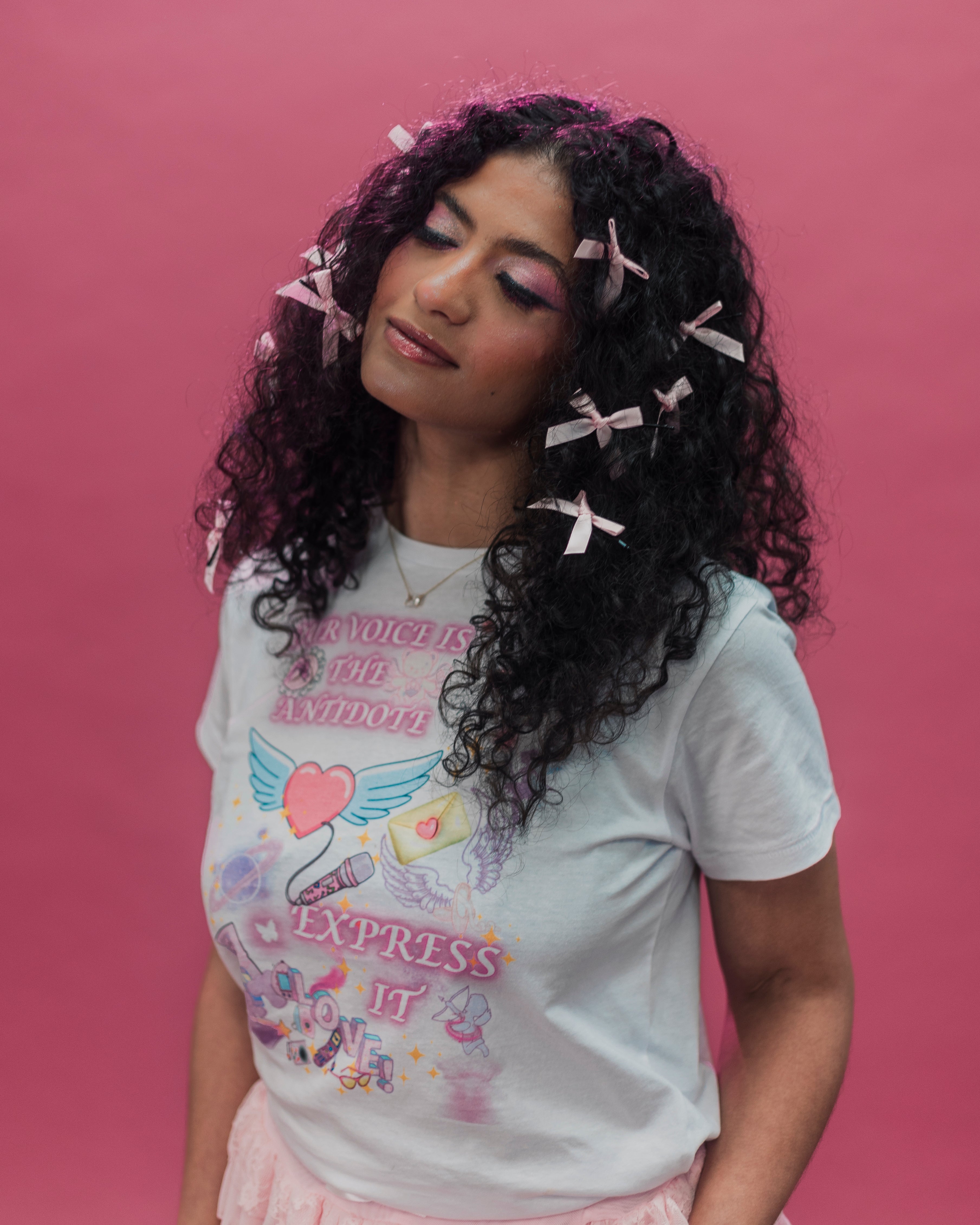 A woman with long, curly black hair adorned with pink ribbon clips, wearing a white graphic t-shirt with a colorful angel design and the text 'OUR VOICE IS THE AN ANTIDOTE' and 'EXPRESS IT'. She is posed against a solid pink background.